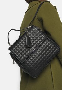 Musta tikattu käsilaukku hopeisilla yksityiskohdilla, jossa on läppä suljenta, säädettävä hihna ja tagi. Raketettu nahka logolla.