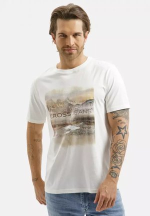 Mężczyzna o średniej długości włosach, ubrany w białą koszulkę z nadrukiem gór i napisem "CROSS JEANS", niebieskie dżinsy oraz widoczne tatuaże na ramionach.