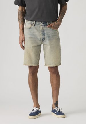 Άνδρας που φορά ξεθωριασμένο κοντό τζιν με φθαρμένες άκρες, σκοτεινό μπλουζάκι, μπλε αθλητικά παπούτσια και εμφανή τατουάζ στα χέρια και το πόδι.