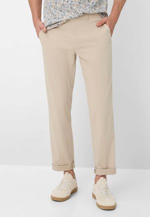 Uomo con chino beige risvoltati, sneakers bianche con suola in gomma e camicia a fantasia floreale, in piedi con le mani nelle tasche.