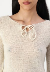 Lyscremet strikket sweater med løs pasform, der har en rund halsudskæring og en dekorativ sløjfe i midten. Blødt, tekstureret stof.