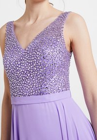 V-kaula-aukollinen iltapuku, jossa kimaltava lilansävyinen yläosa, koristeltu kirkkailla kristalleilla, ja välkkyvä laventelinsävyinen hame. Pehmeä pinta ja elegantti muotoilu.