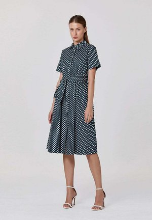 Femme portant une robe bleu marine à pois blancs, manches courtes, ceinture à la taille et sandales blanches à talons hauts, debout devant un fond uni.