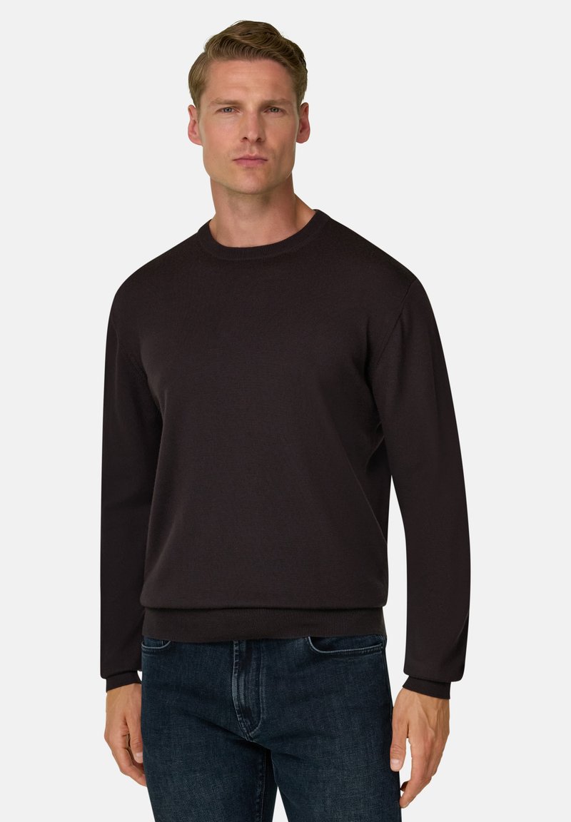 Jeune homme portant un pull noir uni à manches longues et un jean bleu foncé, debout devant un fond clair uni.