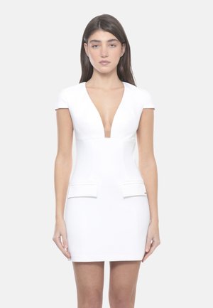 Donna che indossa un vestito bianco aderente a maniche corte con scollo a V profondo e due tasche con patta frontali, in piedi davanti a uno sfondo neutro.