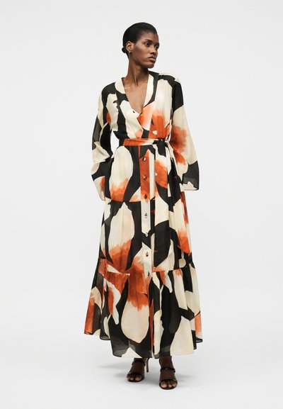 Vrouw die een lange, met knopen gesloten bloemenjurk draagt met zwart, wit en oranje patronen, staand met handen in de zakken en zwarte hakken.