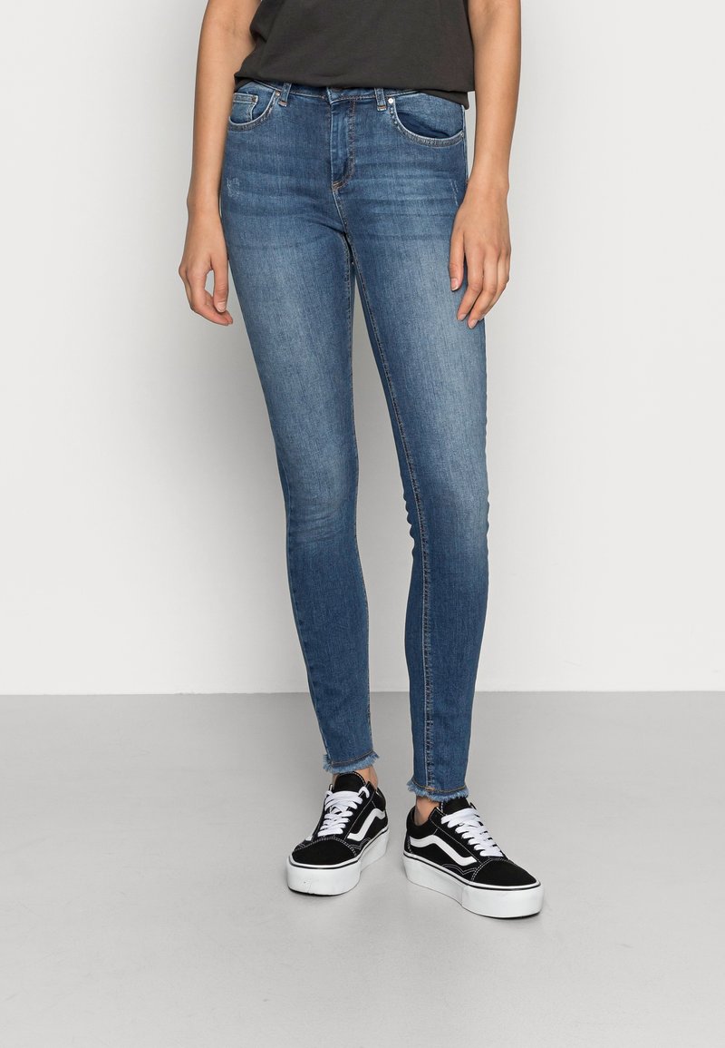 Vaqueros ajustados de denim, de cintura alta, con bolsillos frontales, ligero deshilachado en el dobladillo, combinados con zapatillas negras y blancas.