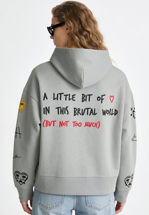 Femme aux cheveux ondulés porte un sweat à capuche gris avec un texte noir et rouge indiquant "A little bit of love in this brutal world (but not too much)" et des patchs graphiques.
