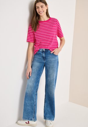 Femme souriante appuyée contre un mur, portant une chemise à rayures roses et blanches, un jean bleu à jambes larges et des baskets blanches avec un accent bordeaux.