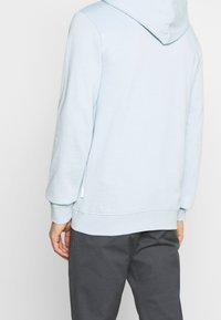 Felpa leggera blu con cappuccio regolabile, polsini e orlo a costine. Dettaglio del tag sul lato, texture morbida e vestibilità comoda. Abbinata a pantaloni grigio scuro.