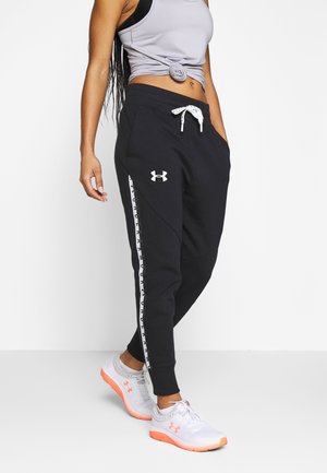 Frau trägt schwarze Under Armour Jogginghosen mit Logo und weißem Kordelzug, graues ärmelloses Oberteil und weiße Sportschuhe mit orangen Sohlen.