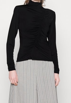 Vrouw draagt een zwarte, lange mouwen, gerimpelde coltrui en een lichtgrijze, hoge taille wijde broek met dunne verticale strepen.