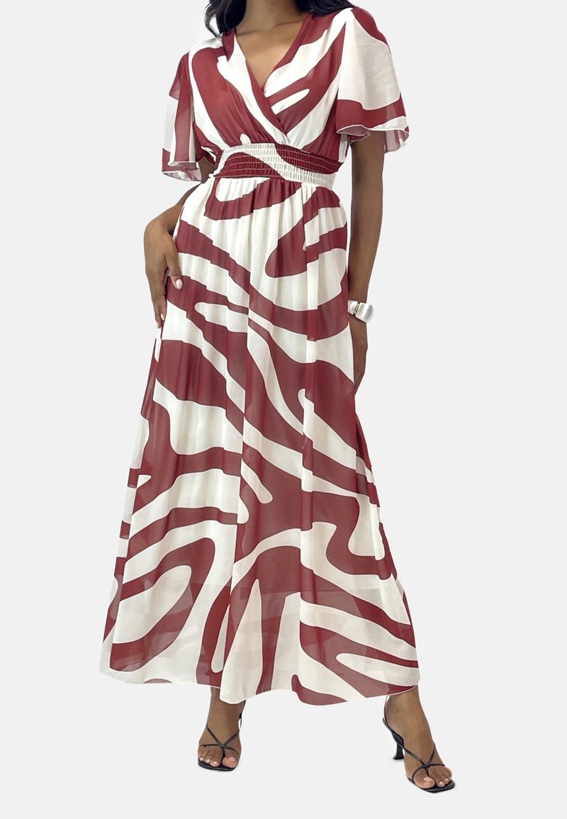Femme portant une longue robe fluide avec de larges rayures abstraites bordeaux et blanches, un décolleté en V profond, des manches flottantes et des talons noirs à brides.