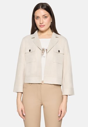 Veste courte beige en tissu doux avec un col, deux poches à rabat et des boutons décoratifs. Portée sur un T-shirt blanc et un pantalon beige.
