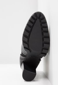 Bottines noires à talons hauts avec une semelle en caoutchouc texturée, présentant un large orteil et un talon courbé pour plus de stabilité. La tige a des accents en cuir.