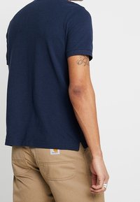 Navyblå bomulls-t-shirt med korta ärmar, rundad krage och rak nederkant. Synlig tatuering på armen, bärs med khakifärgade byxor.