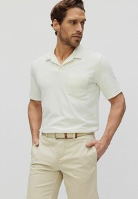 Kortärmad, ljusbeige polo med ficka, med en avslappnad krage och mjuk textur, tillsammans med ljusa byxor och ett bälte.