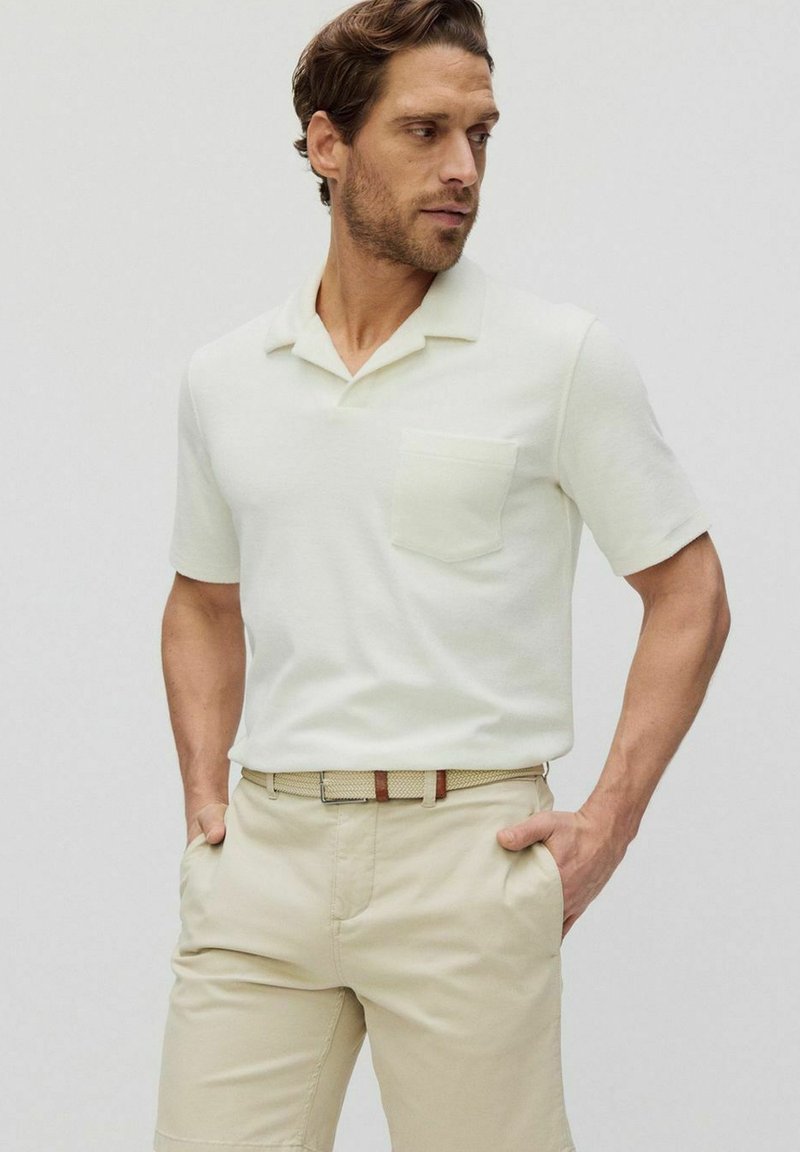 Kortärmad, ljusbeige polo med ficka, med en avslappnad krage och mjuk textur, tillsammans med ljusa byxor och ett bälte.