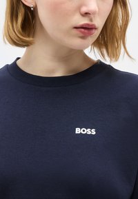 Γυναίκα με κοντά ανοιχτά καστανά μαλλιά φοράει μπλε πουκάμισο με το λευκό λογότυπο "BOSS" στο στήθος και ασημένιο κρίκο στο αριστερό αυτί.