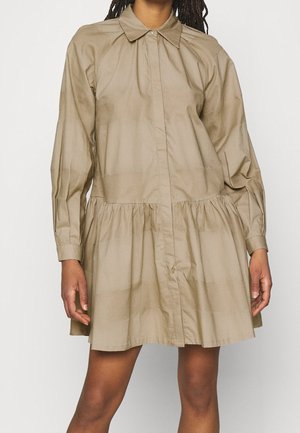 Femme portant une robe beige ample à manches longues, col et taille froncée, debout devant un fond uni.