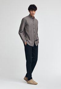 Brązowa koszula z długim rękawem i zapięciem na guziki o subtelnym wzorze, zestawiona z czarnymi spodniami i beżowymi sneakersami. Koszula ma detal w postaci kieszeni.