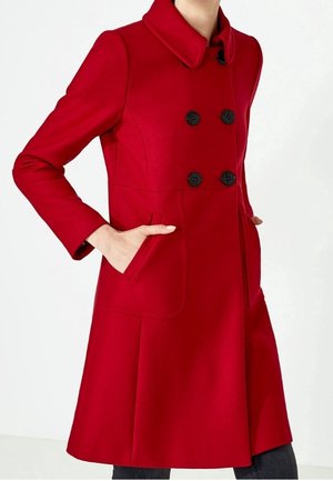 Kobieta w dopasowanym czerwonym płaszczu z czarnymi dwurzędowymi guzikami, z rękami w bocznych kieszeniach na białym tle.