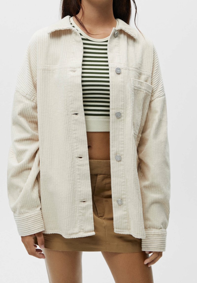 Lys beige corduroy oversized skjortejakke med lodrette ribber, frontknapper og en enkelt lomme. Sat sammen med en grønstribet crop top.