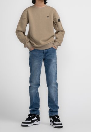 Sweat-shirt beige à manches longues et col rond, associé à un jean bleu. Chaussures : baskets noires et blanches à motifs.