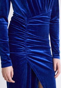 Vestido de veludo azul royal com mangas longas, apresentando um design franzido na cintura e uma fenda até a coxa de um lado. Textura suave.