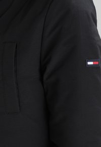 Manche de veste noire avec un petit patch rectangulaire présentant un logo rouge, blanc et bleu sur le bras supérieur.