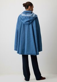 Lys blå oversized denimfrakke med hætte, der har krave og en længere bagkant. Stylet over mørke bukser og brune sko.