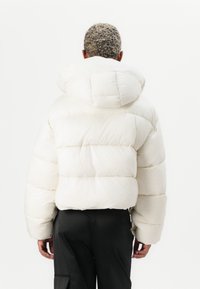 Crème pufferjack, voorzien van een capuchon, een boxy ontwerp, een gewatteerde textuur en brede, puffy mouwen. Gedragen over zwarte broeken.