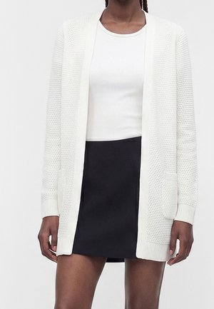 Kvinde iført en hvid struktureret cardigan med åben front over en hvid skjorte og sort nederdel, stående mod en ensfarvet baggrund.