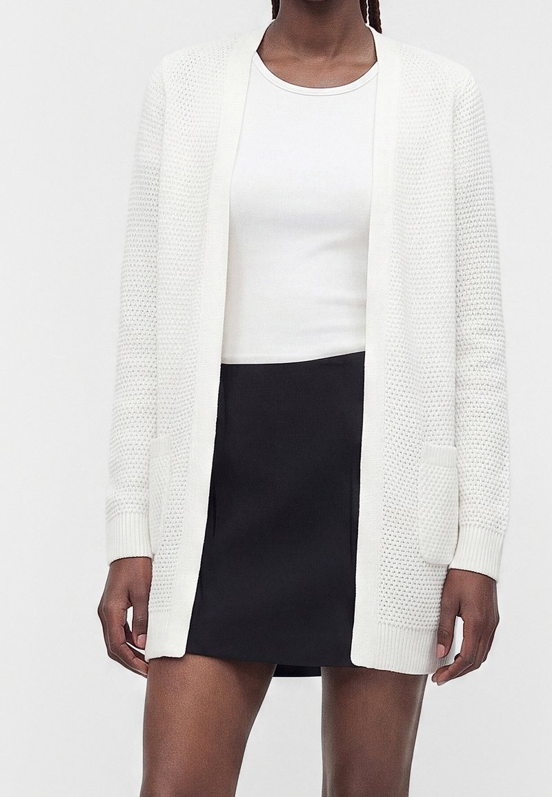 Femme portant un cardigan blanc texturé ouvert sur le devant, par-dessus une chemise blanche et une jupe noire, debout devant un fond uni.