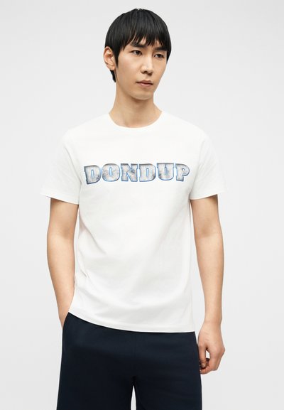 Νέος άνδρας που φοράει λευκό μπλουζάκι με το κείμενο «DONDUP» σε μπλε-γκρι εκτύπωση και σκούρο παντελόνι, στέκεται με το ένα χέρι στην τσέπη.