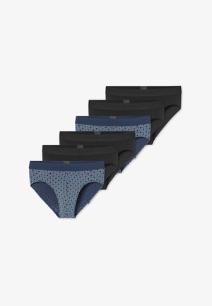 Hombre musculoso con cabello corto, vistiendo calzoncillos azules con estampado, de pie con los brazos cruzados, mirando hacia un lado sobre un fondo liso.