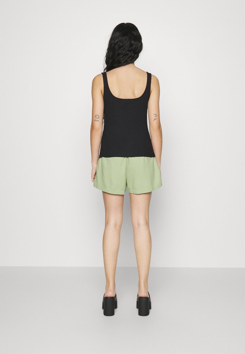 Femme debout de dos, vêtue d'un haut noir sans manches, d'un short vert clair et de chaussures compensées noires, avec des tatouages sur les deux bras supérieurs.