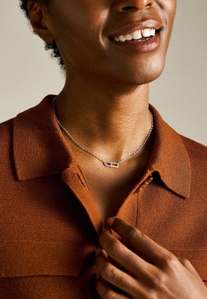 Collana in argento con un pendente a catena, indossata sopra una camicia color marrone texturizzata. Il metallo liscio contrasta con la texture del tessuto.