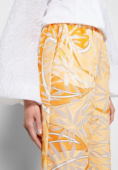 Mangas longas brancas texturizadas combinadas com calças amarelas estampadas com desenhos de folhas em branco e cinza claro, apresentando uma palete de cores vibrante.