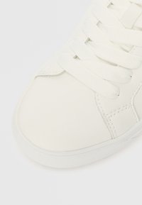 Λευκό αθλητικό παπούτσι με λεί�ο δερμάτινο επάνω μέρος, στρογγυλή μύτη και επίπεδες λευκές κορδόνες. Η σόλα είναι υφασμένη με σχέδιο για καλύτερη πρόσφυση.