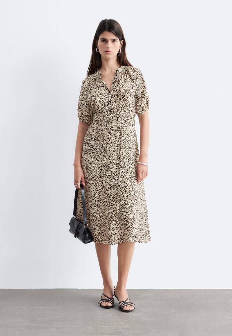 Woman standing against a plain wall, wearing a floral-patterned midi dress, black sandals, holding a small black handbag, neutral expression.
