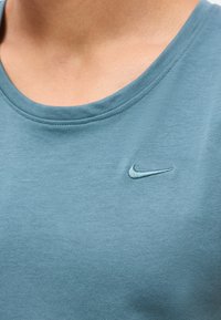 Κοντινό πλάνο ατόμου που φοράει μπλε-πράσινο μπλουζάκι Nike με μικρό κεντημένο λογότυπο Nike στο αριστερό στήθος.