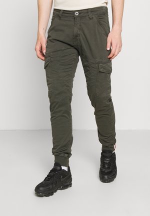 Persona con pantalones cargo verde oliva y zapatillas negras de pie contra un fondo claro y liso.