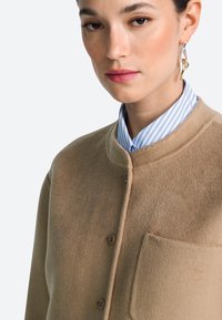 Beige jas van een wollen mix met een ronde kraag, knopen aan de voorkant en een borstzak. Draagt over een blauw-wit gestreept overhemd met kraag.