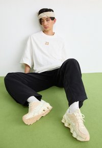 Camisa branca de manga curta com riscas verticais e detalhe "23", calças soltas pretas e ténis de cor clara com sola texturada.