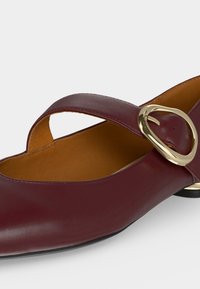 Chaussure plate en cuir bordeaux avec une large bride sur le dessus, maintenue par une grande boucle ovale dorée et une doublure intérieure couleur caramel.