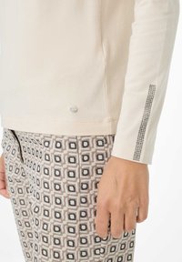 Hand an der Seite ruhend, trägt ein beige langärmliges Oberteil mit einem schmalen Strassstreifen am Ärmel und gemusterte Hose mit geometrischem Quadratdesign.