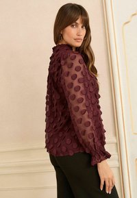 Blusa trasparente color bordeaux con pois testurizzati, scollo ruffled e polsini smockati. Maniche lunghe e vestibilità comoda, indossata con pantaloni neri.