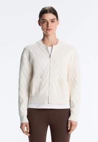 Femme portant un cardigan matelassé blanc à fermeture éclair avec deux poches avant, accompagné d'un pantalon marron et d'une chemise blanche en dessous.