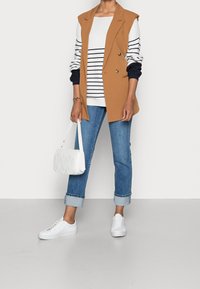 Ärmelloser blauer Blazer über einem gestreiften weißen und marineblauen Pullover, kombiniert mit hochgekrempelten blauen Jeans und weißen Sneakers. Eine kleine gequiltete weiße Handtasche.